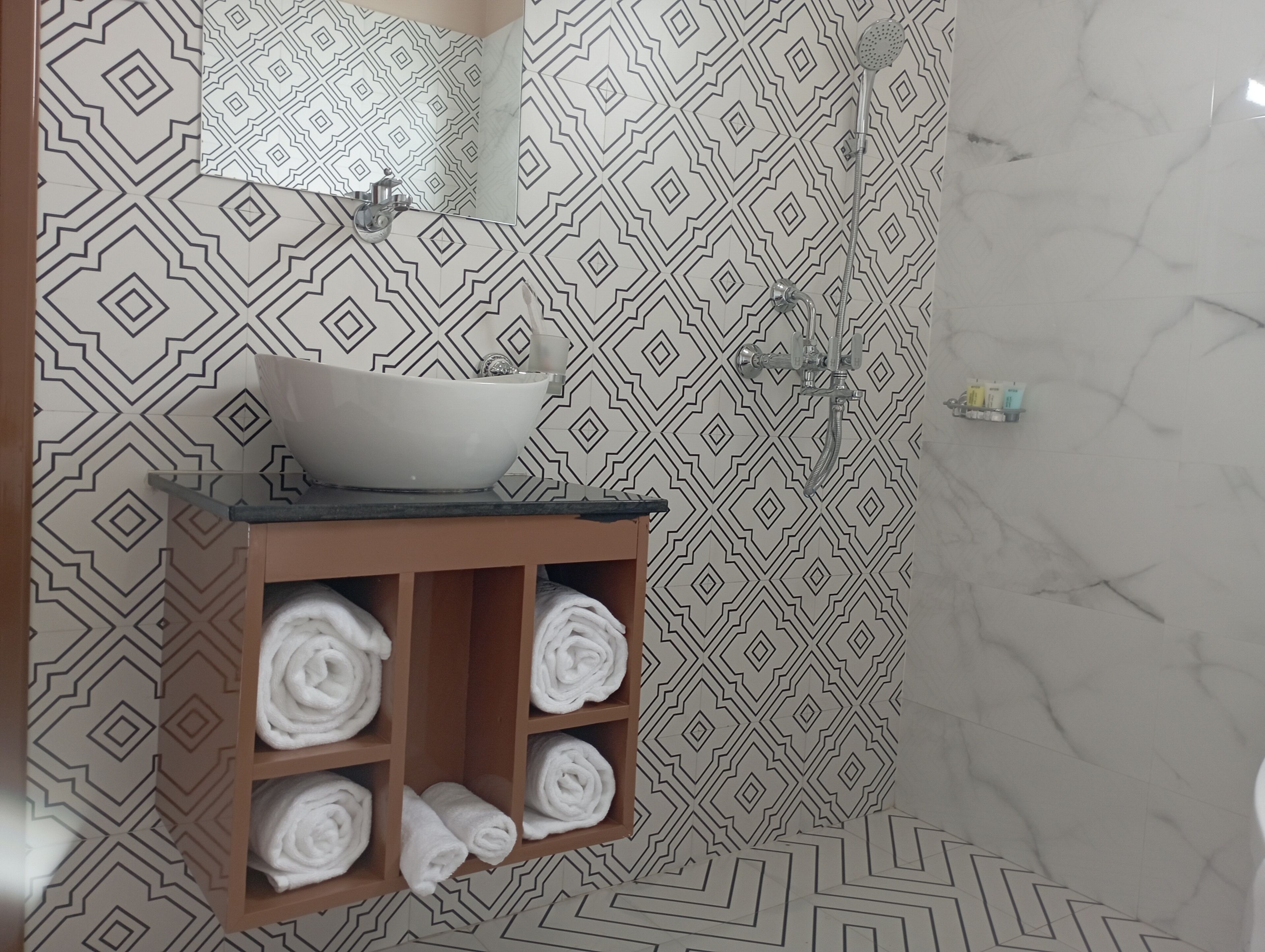 A bathroom with a geometric patterned wall, a white vessel sink, and a wooden shelf with towels.