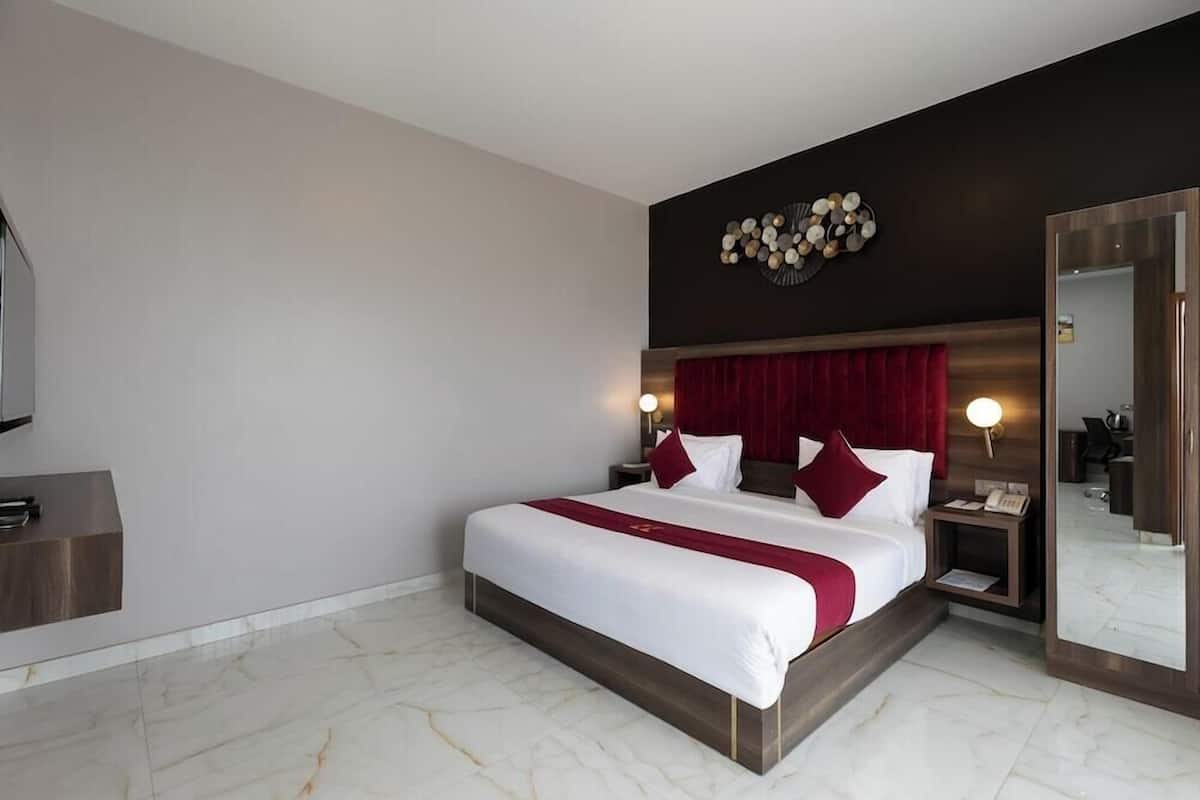 A modern hotel room with a large bed, a dark headboard, a wall-mounted clock, a bedside table with a lamp, and a large mirror.