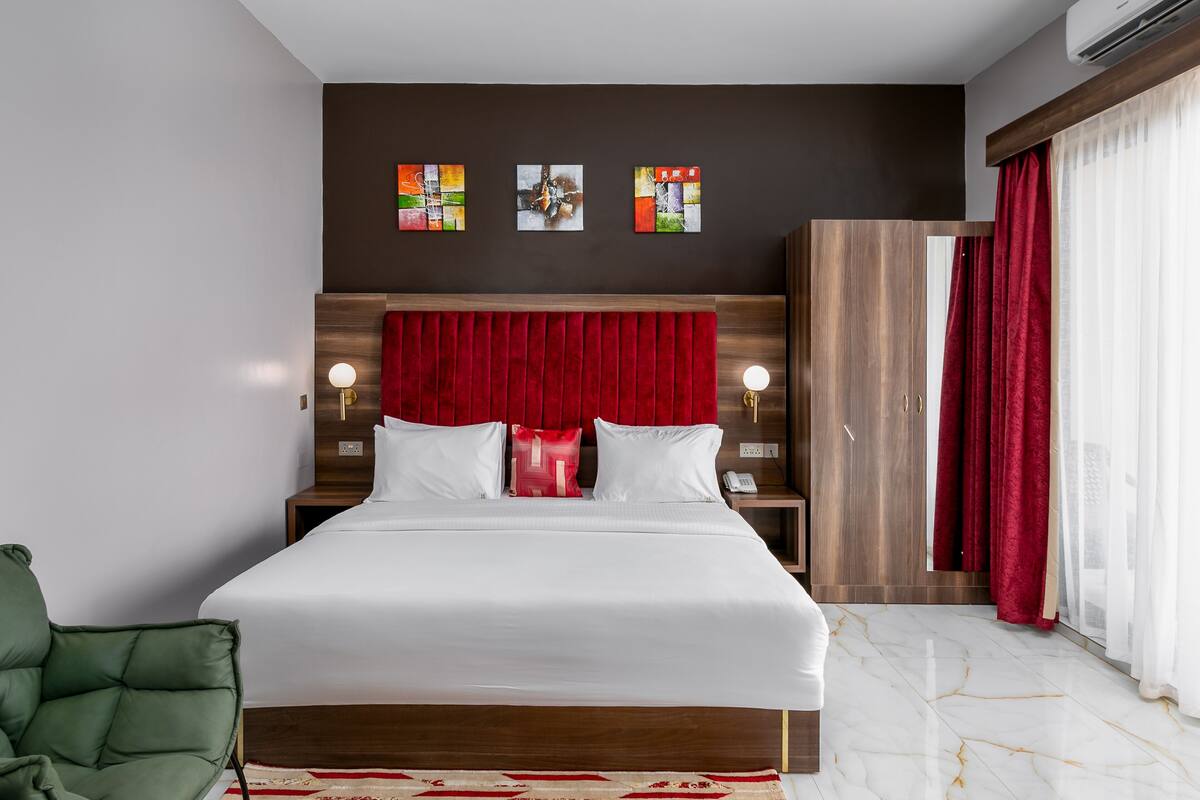 A hotel room with a large bed, a green chair, a wooden wardrobe, and artwork on the wall.