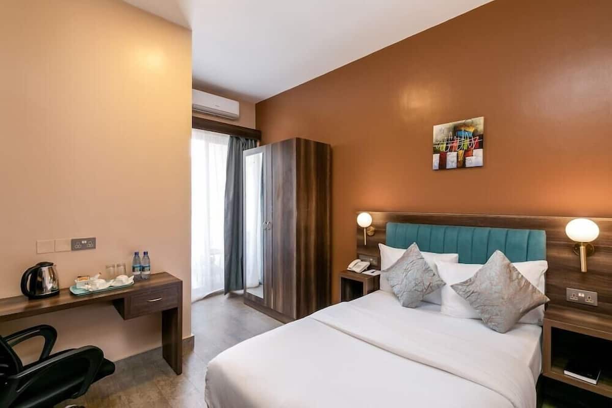 A hotel room with a bed, bedside tables, a desk, a chair, a wardrobe, and a painting on the wall.