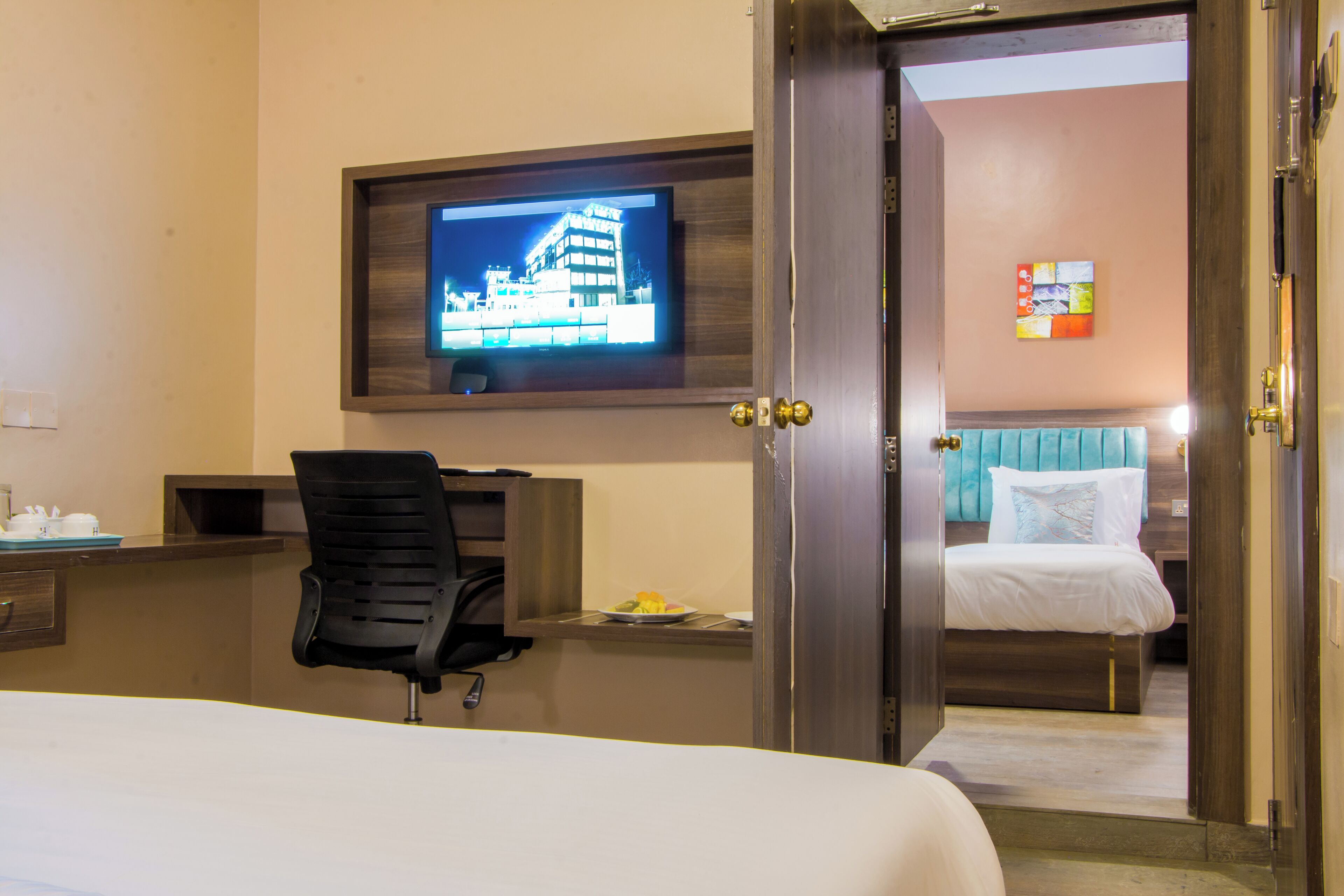 A hotel room with a bed, a desk with a chair, a television mounted on the wall, and a door leading to another room.