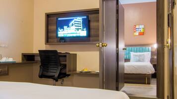 A hotel room with a bed, a desk with a chair, a television mounted on the wall, and a door leading to another room.