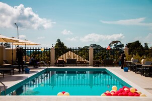 Una piscina con sillas de descanso y sombrillas, rodeada por una valla y árboles.