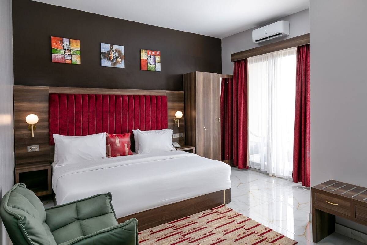 A modern hotel room with a large bed, red curtains, a green armchair, and artwork on the wall.