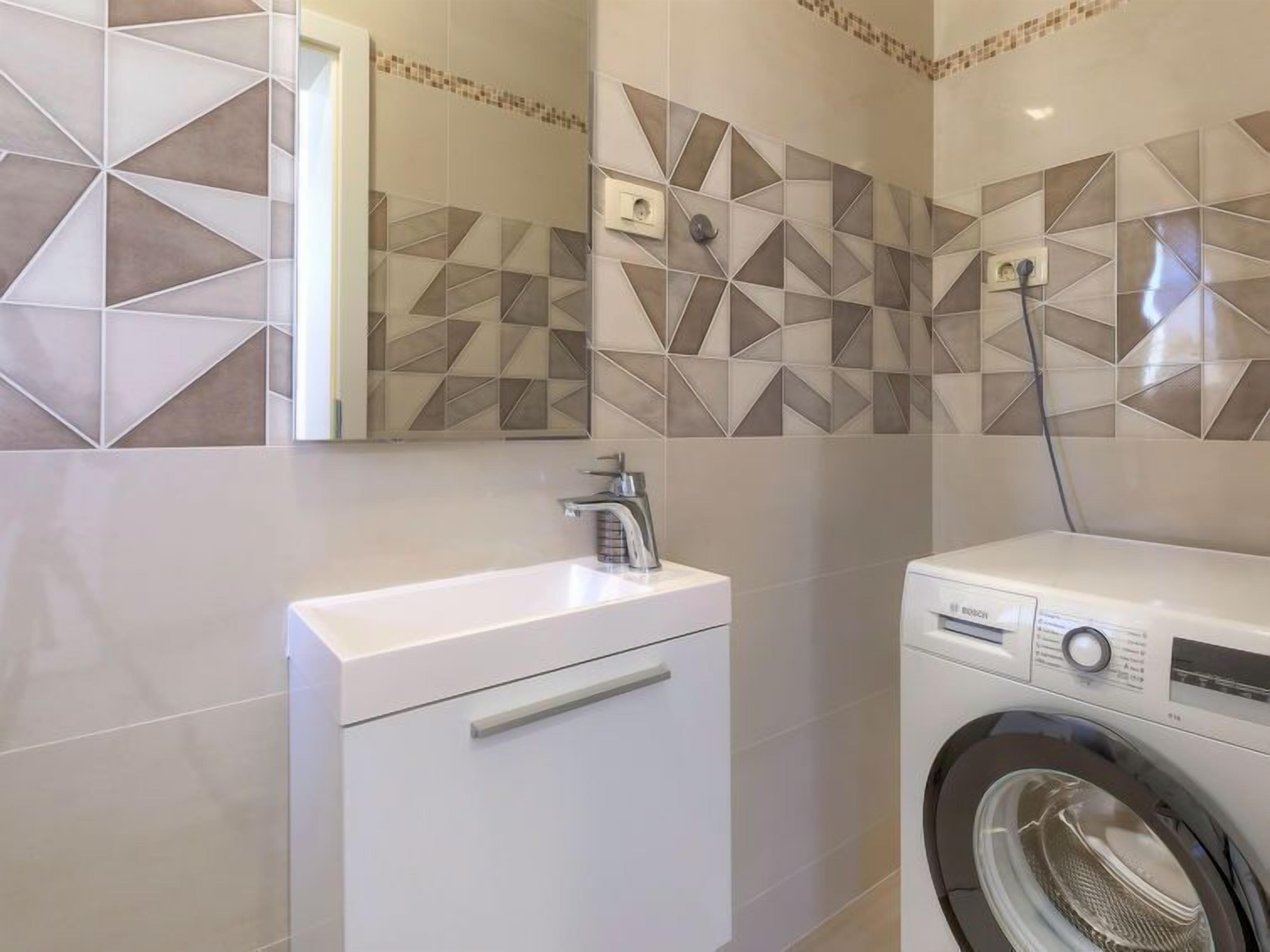 A modern bathroom with a white sink, a Bosch washing machine, and geometric patterned tiles.
