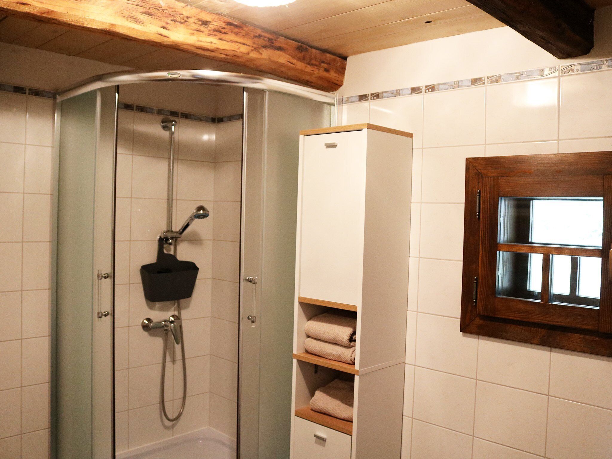 Een douche met glazen deur, een houten plafond en een witte betegelde muur met een raam met houten kozijn.