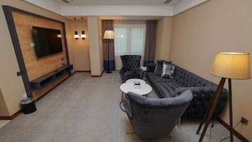 Una habitación de hotel moderna con un televisor de pantalla plana montado en un panel de madera, un sofá gris, una pequeña mesa redonda y una lámpara de pie.