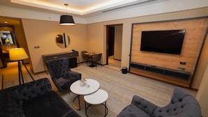 Et moderne hotelværelse med et stort fladskærms-tv monteret på en trævæg, et rundt spisebord med stole og en siddeplads med polstrede sofaer og et lille sofabord.