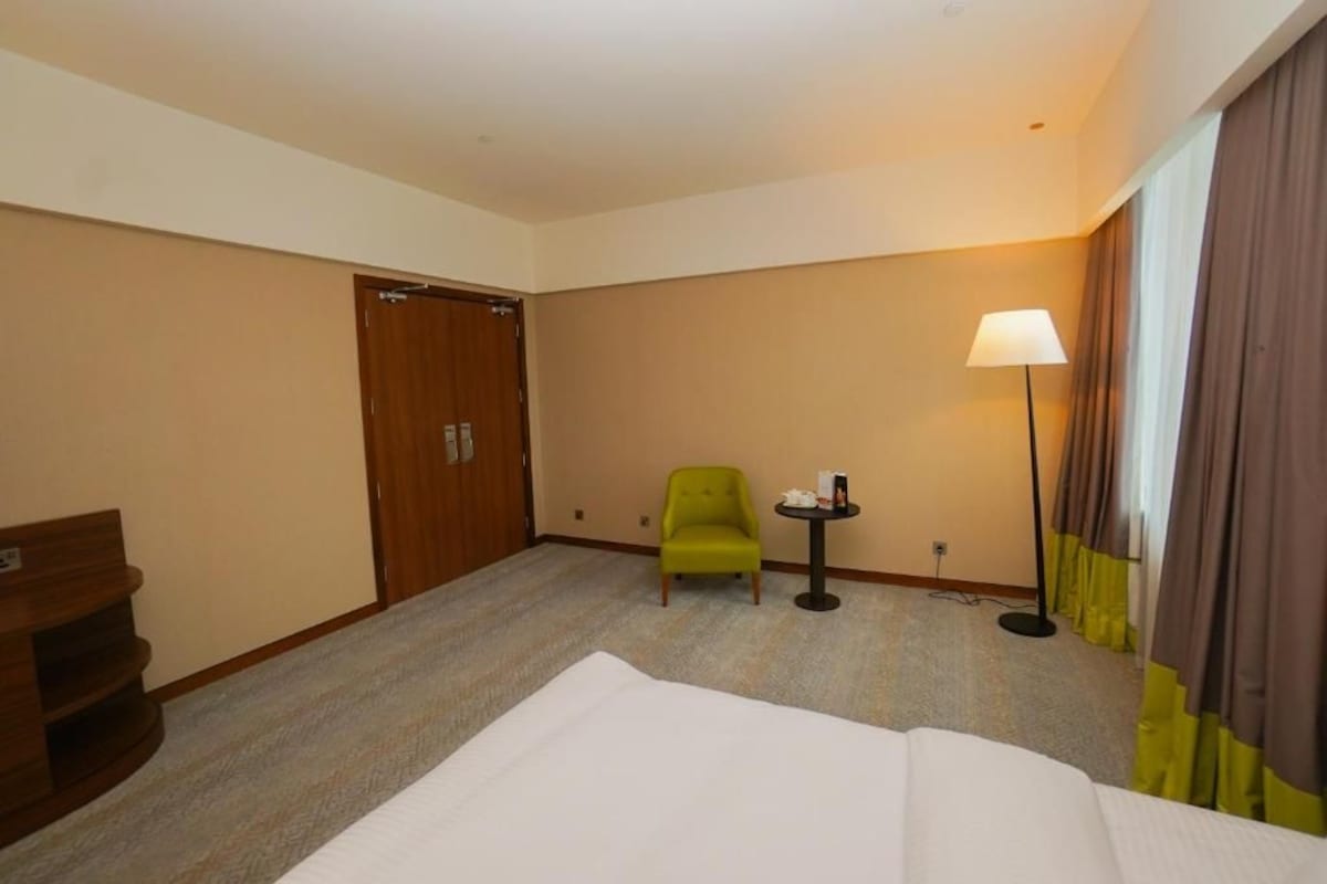 A hotel room with a bed, a green armchair, a small table with a lamp, and a wardrobe.