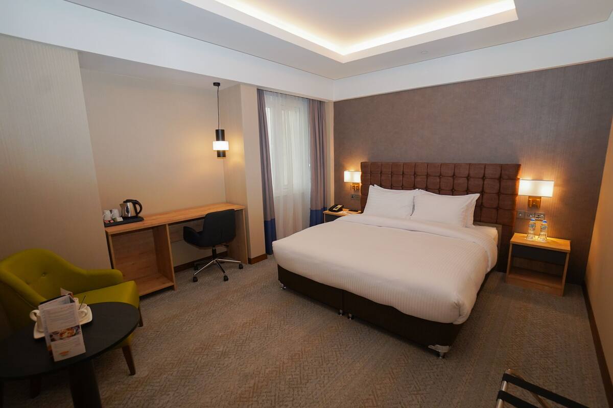 A hotel room with a large bed, a desk with a chair, a small table with a chair, and a nightstand with a lamp.