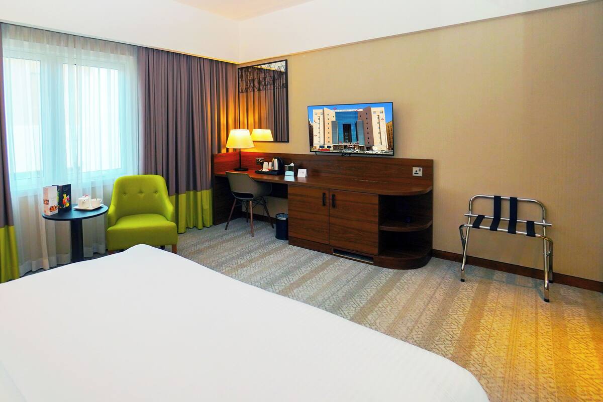 A hotel room with a bed, a desk, a chair, a lamp, a mirror, and a window with curtains.
