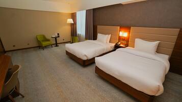 A hotel room with two beds, a desk, a chair, and a lamp.