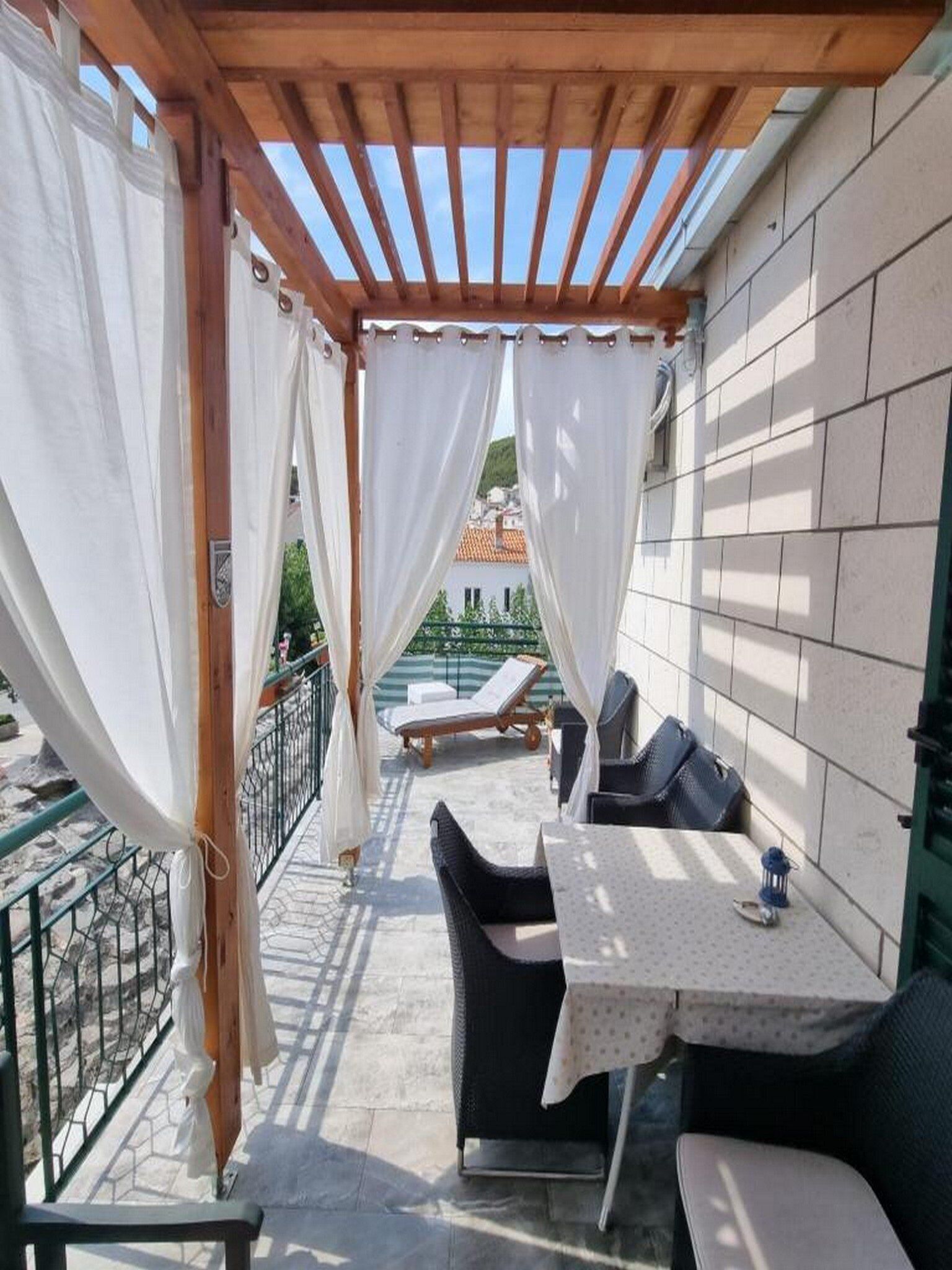 Uma área de estar coberta ao ar livre, com mesa e cadeiras, vista para um edifício ao fundo e um corrimão em um dos lados.