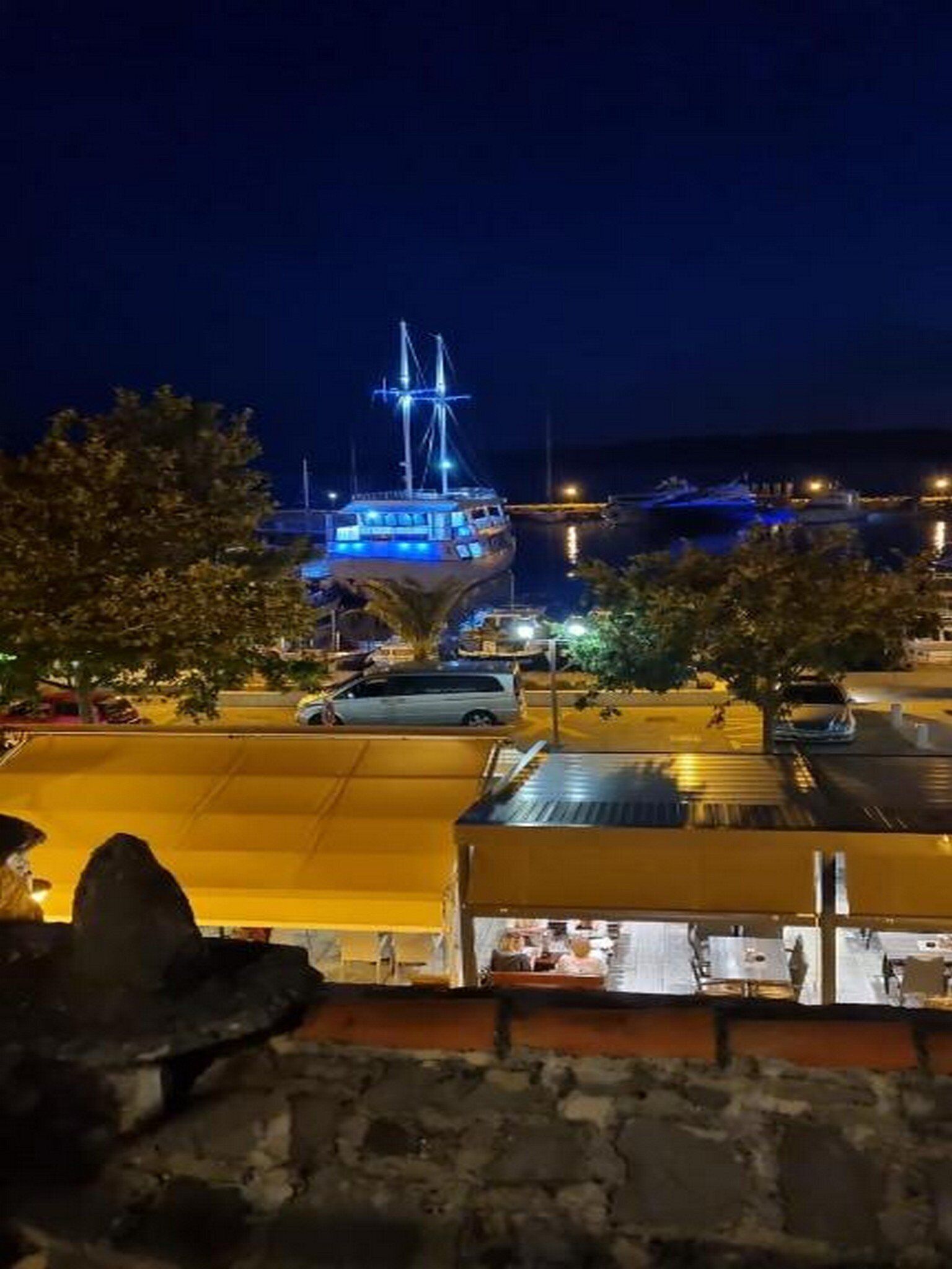 Een nachtzicht van een waterkant met een groot, verlicht schip aangemeerd aan een steiger, omringd door geparkeerde auto's en gebouwen met zichtbare ramen.