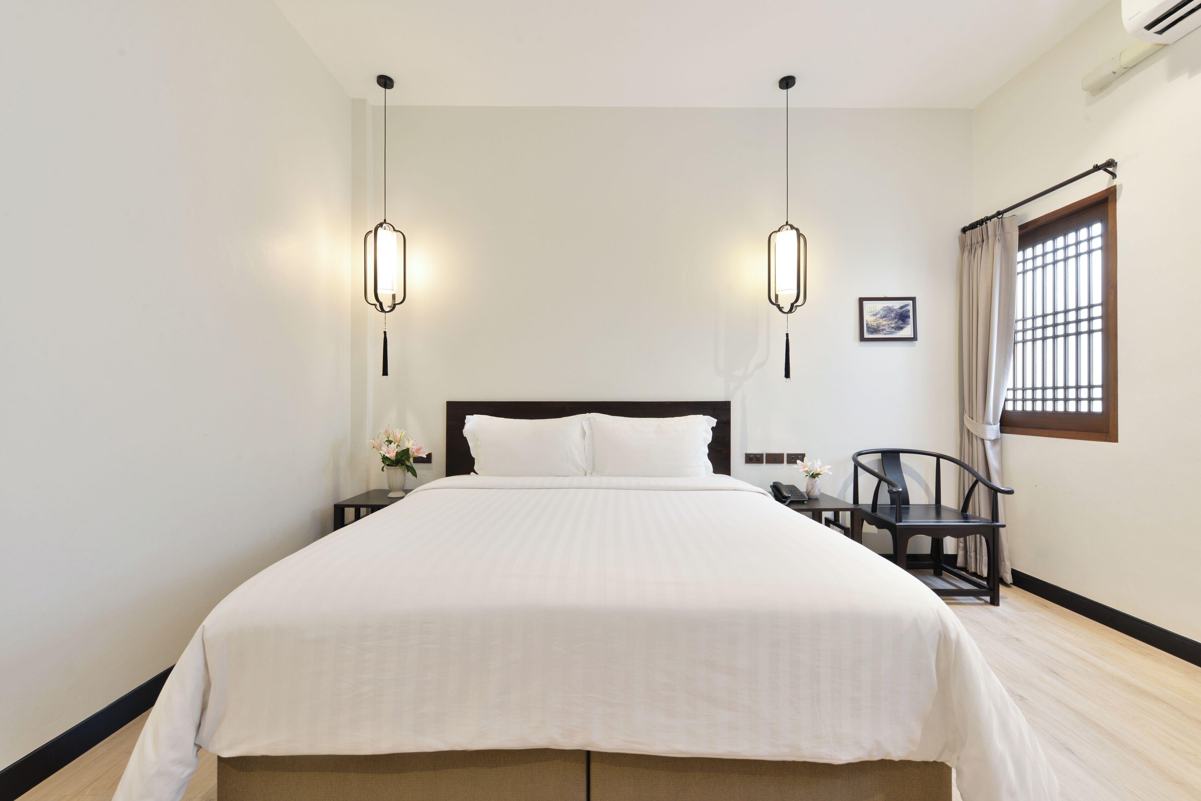 A neatly made bed with a white bedspread, two bedside tables with lamps, a chair, and a window with curtains.