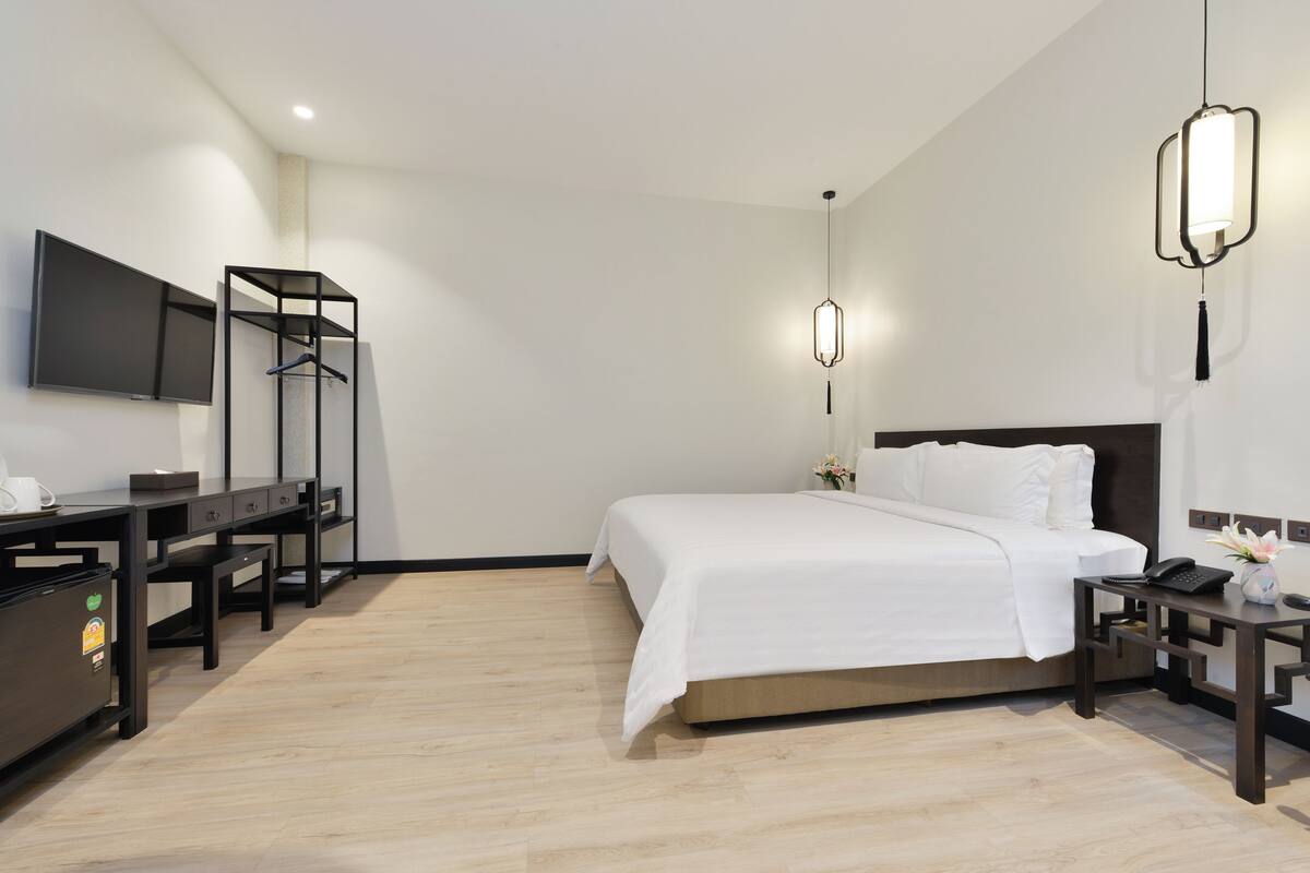 A modern hotel room with a large bed, a desk with a chair, a TV mounted on the wall, and a hanging light fixture.