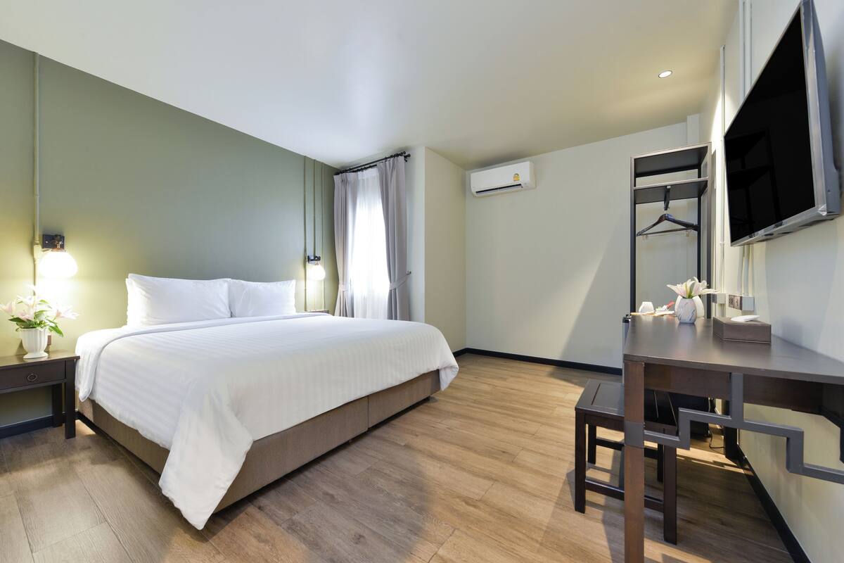 A hotel room with a bed, bedside table, lamp, air conditioning unit, and a desk with a chair.