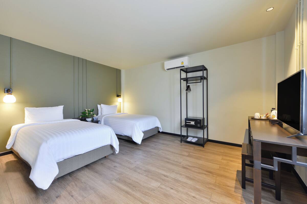 A hotel room with two beds, a desk, a TV, and an air conditioning unit.