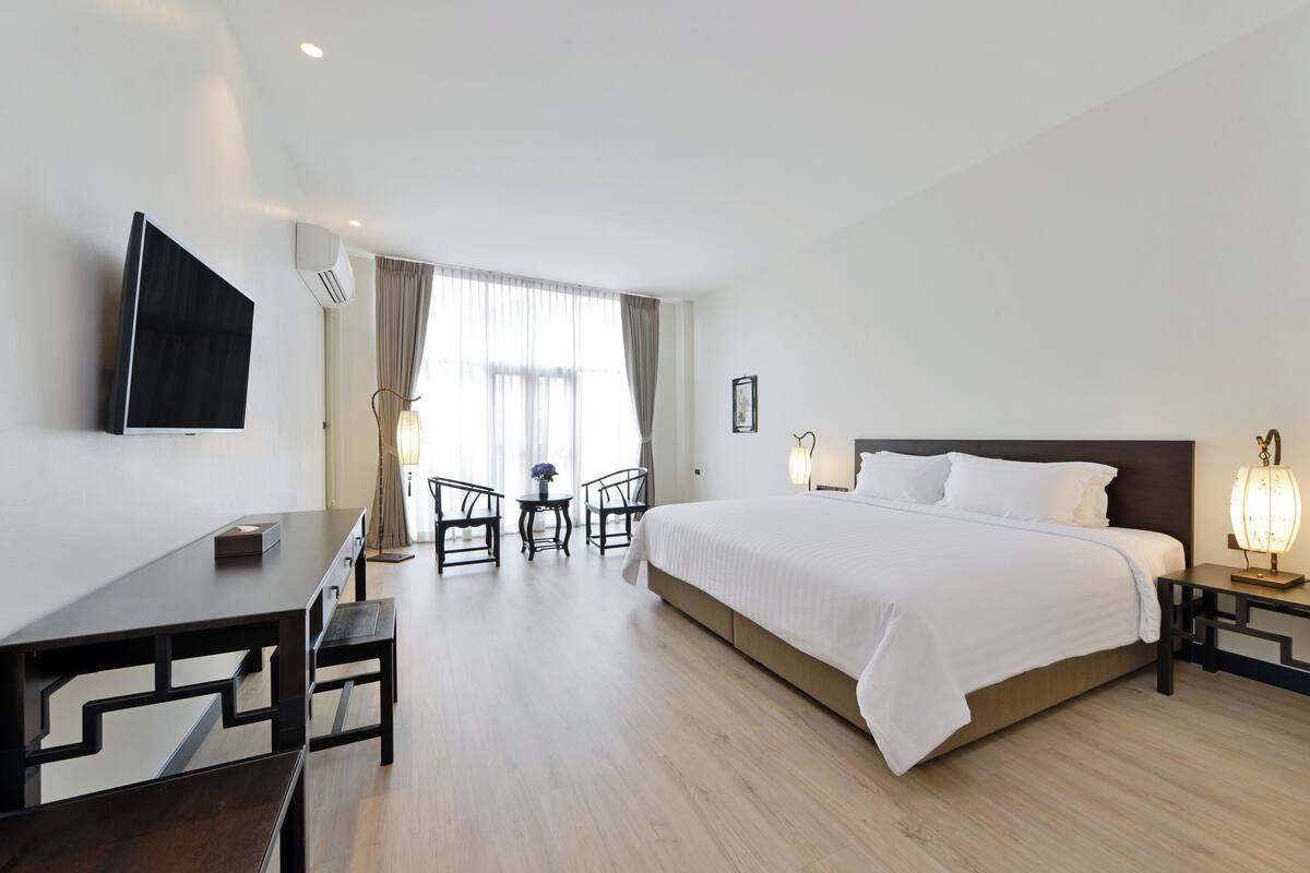 A hotel room with a large bed, a desk, two chairs, a TV, and a window with curtains.
