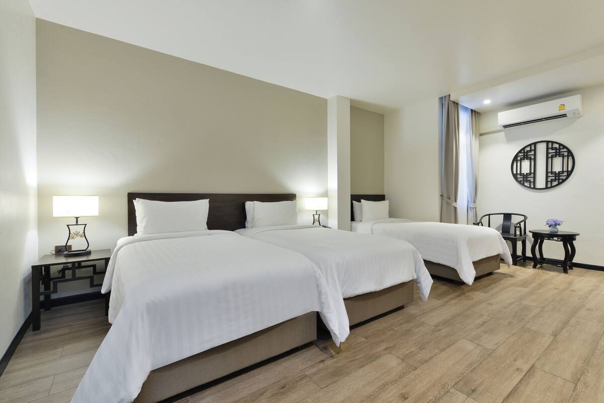 A hotel room with two beds, a nightstand with a lamp, a small table with a vase, and an air conditioning unit.
