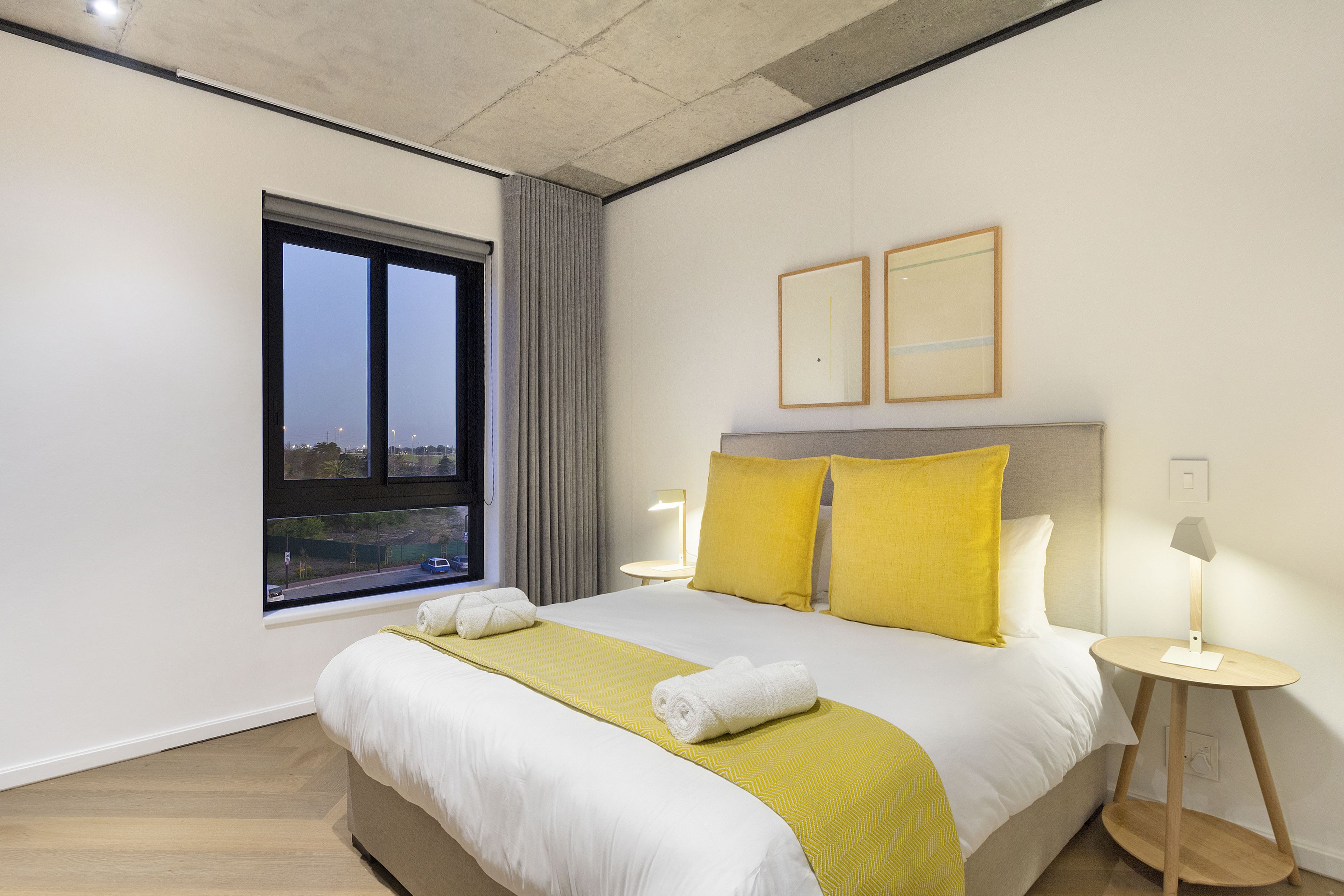A hotel room with a large bed, two bedside tables with lamps, a window with curtains, and two framed pictures on the wall.