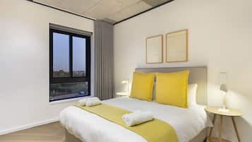 Une chambre d’hôtel avec un grand lit, deux tables de chevet avec des lampes, une fenêtre avec des rideaux et deux tableaux encadrés au mur.