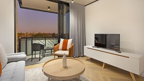 A modern living room with a sofa, a round coffee table, a TV on a white cabinet, and a balcony with a view of the city at dusk. - Bridgewater View 502 (Cape Town)