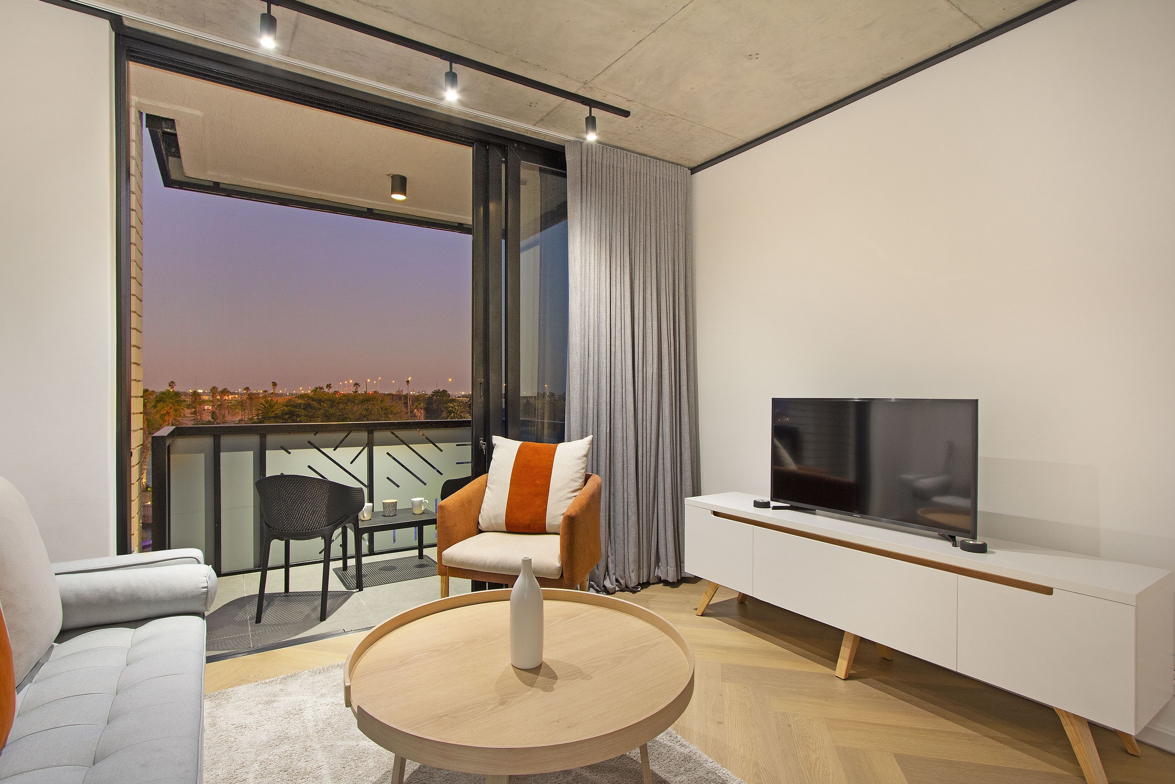 Un salon moderne avec un canapé, une table basse ronde, un téléviseur posé sur un meuble blanc et un balcon offrant une vue sur la ville au crépuscule.