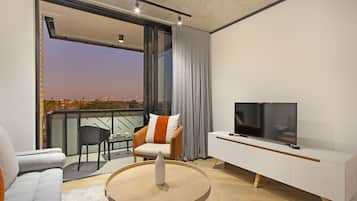 Un salon moderne avec un canapé, une table basse ronde, un téléviseur posé sur un meuble blanc et un balcon offrant une vue sur la ville au crépuscule.