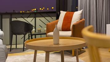 Un salon moderne avec une table ronde en bois, un vase blanc, un canapé agrémenté d’un coussin à rayures et une vue sur les lumières de la ville, visible à travers une fenêtre.