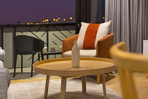 A modern living room with a round wooden table, a sofa with a striped pillow, a black chair, and a view of the city lights through a window.