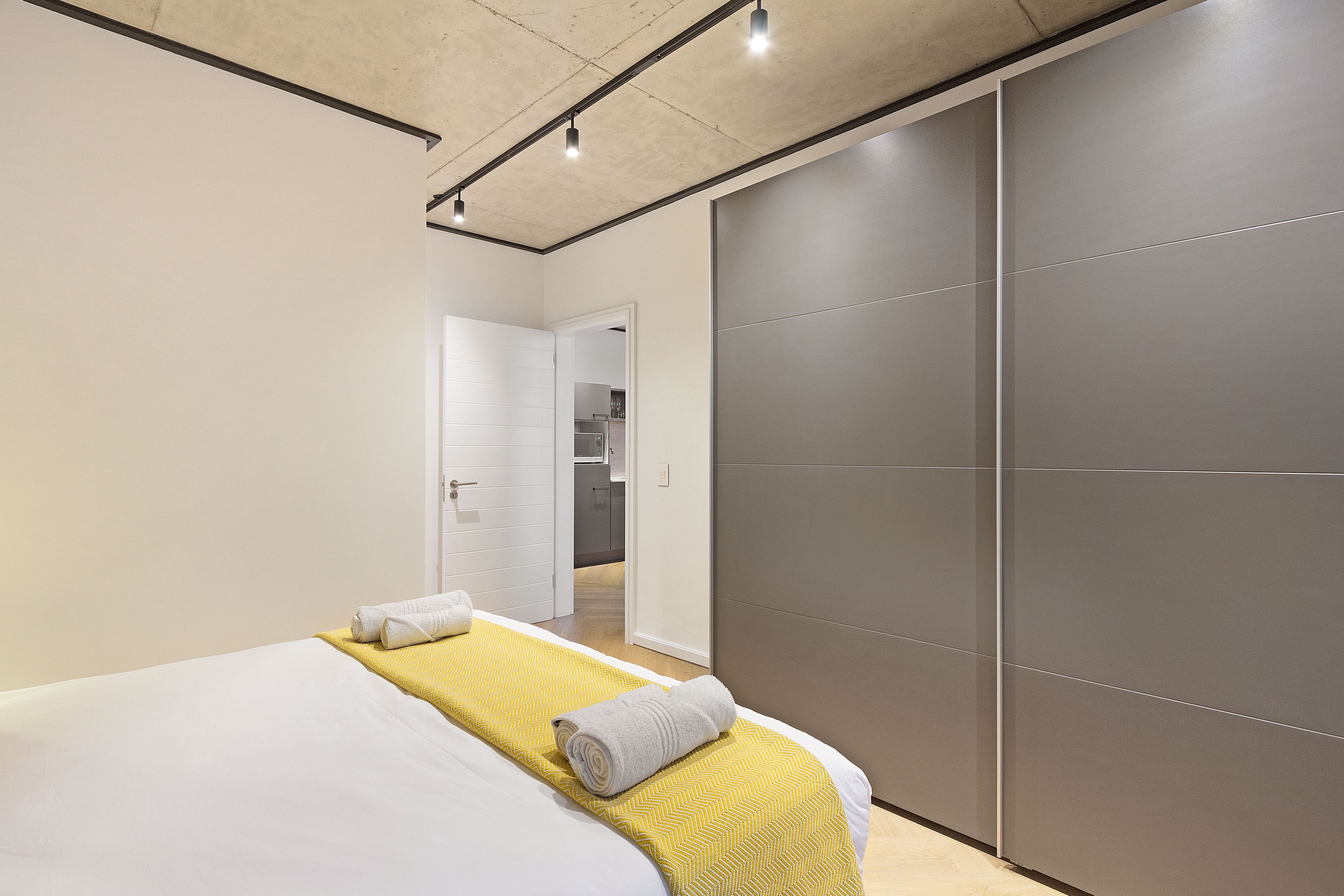 A modern hotel room with a bed, a yellow bedspread, a grey towel, and a sliding door to the bathroom.