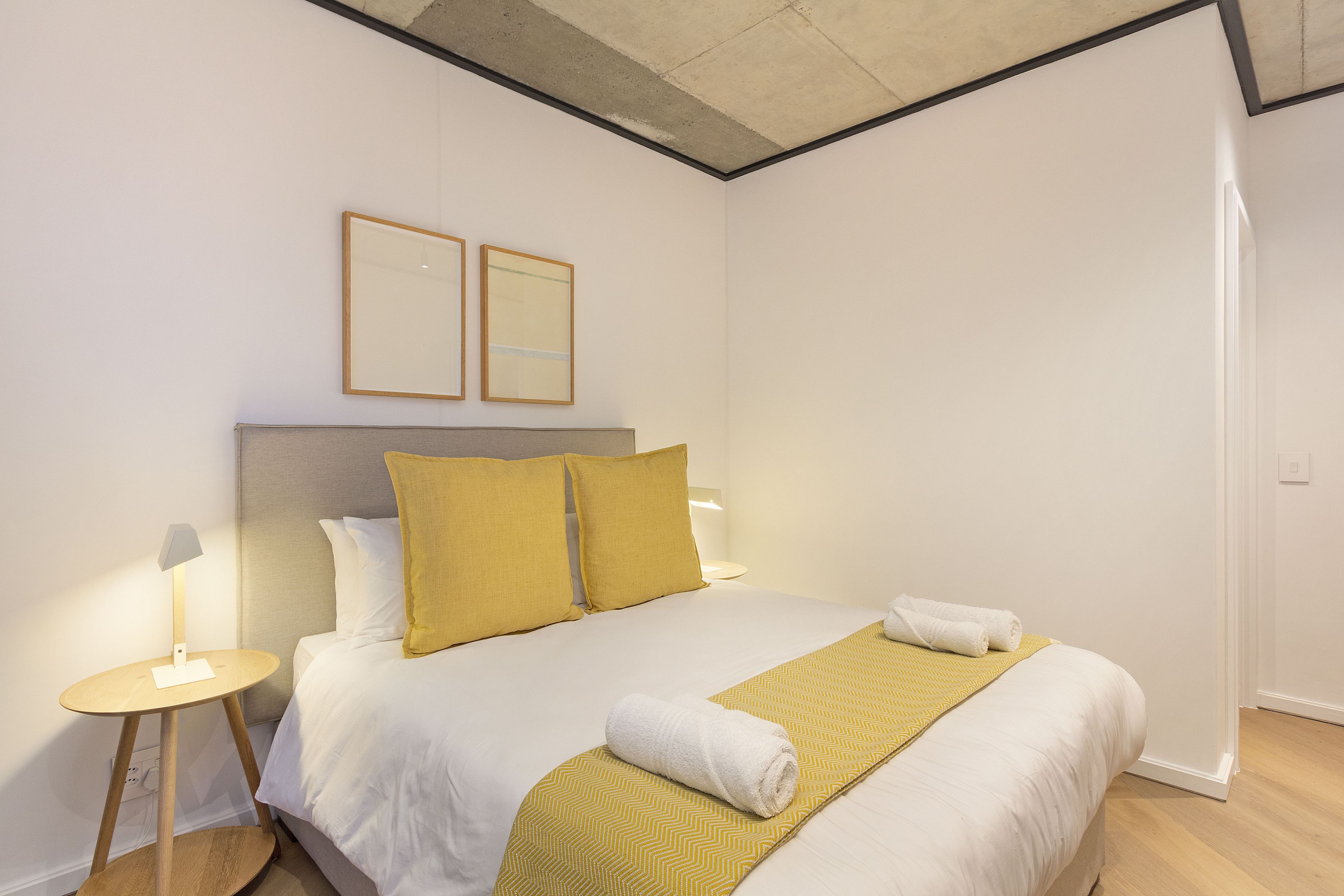 Ein ordentlich gemachtes Bett mit einer weißen und gelben Tagesdecke, ein aufgerolltes weißes Handtuch und zwei gerahmte Bilder an der Wand darüber.