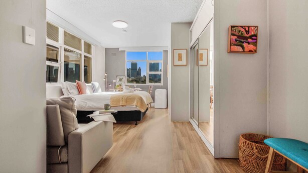 A modern hotel room with a large bed, a sofa, a small table, and a view of the city through large windows. - Location and lifestyle in a cute luxe studio with stunning city views (Woolloomooloo)