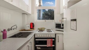 A compact kitchen with white cabinets, a built-in oven, a microwave, and a window with a view of palm trees. - Location and lifestyle in a cute luxe studio with stunning city views (Woolloomooloo)