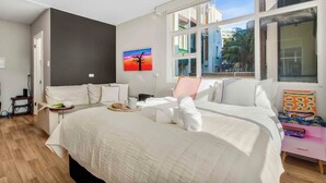 A hotel room with a large bed, a window with a view of buildings, a painting on the wall, and a small table with a bowl and a cup. - Location and lifestyle in a cute luxe studio with stunning city views (Woolloomooloo)