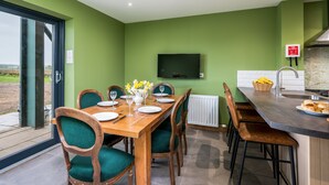 Un coin repas avec une table en bois dressée pour quatre personnes, des chaises à l’assise rembourrée de tissu vert, un vase rempli de fleurs et une vue sur l’extérieur grâce à une porte vitrée.