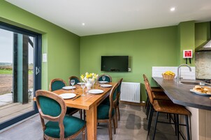 Un comedor con una mesa de madera dispuesta para cuatro comensales, sillas tapizadas de verde, un jarrón con flores y vistas al exterior a través de un ventanal.