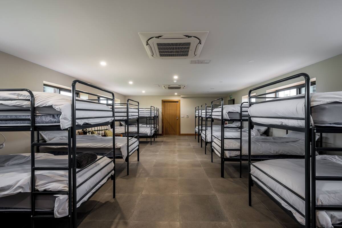 A dormitory room with bunk beds arranged in two rows, a wooden door, and a ceiling vent.
