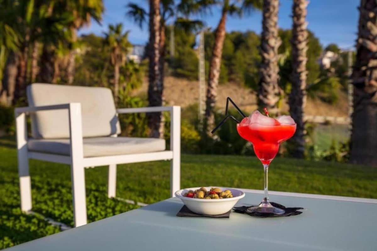 Una mesa con una copa de martini y una ensalada de frutas, una silla blanca, y vistas a palmeras.