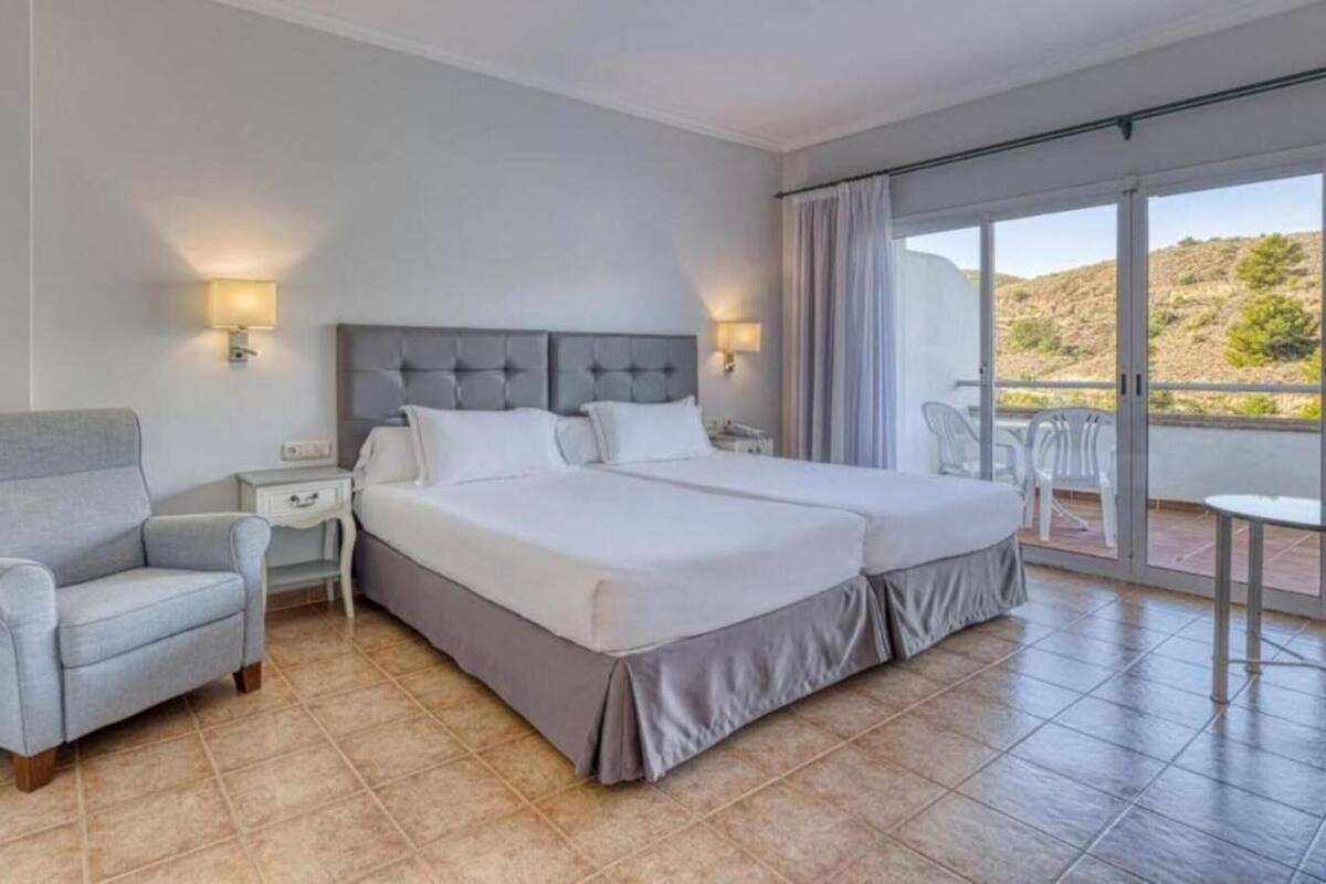 Habitación de hotel con una cama grande, una silla, una mesita de noche y un balcón con vistas a las colinas.
