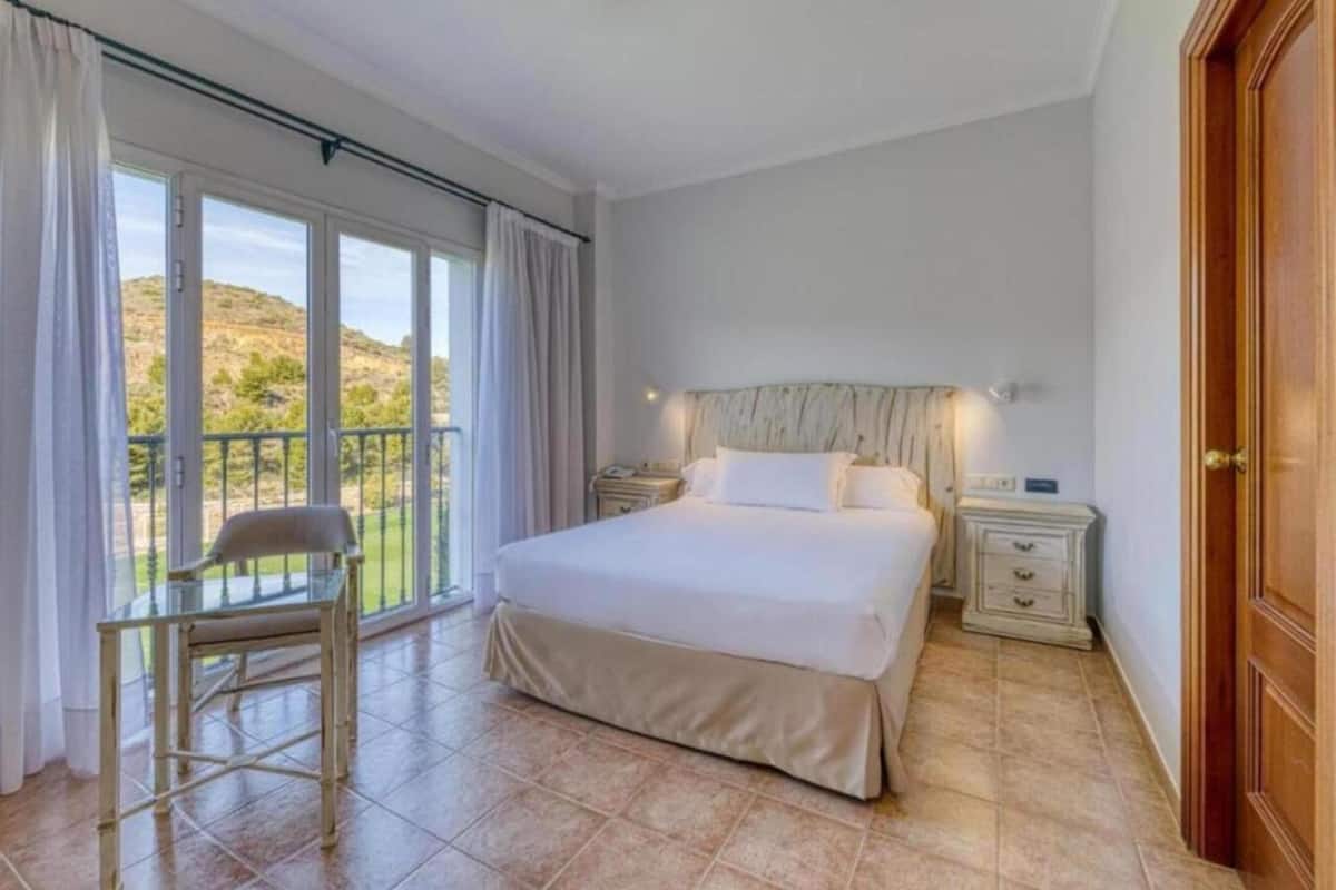 Un dormitorio con una cama grande, una silla, una mesita de noche y vistas al exterior.