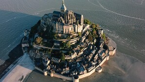 An aerial view of a historic walled city with a prominent church tower, surrounded by water and featuring a mix of residential and fortified structures. - Le Petit Gîte du Mont, gîte at the gates of Mont Saint Michel (Beauvoir)