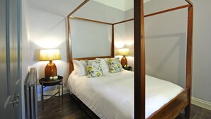 Ein Bett mit Baldachin, dessen Gestell aus Holz besteht, zwei Nachttische mit Lampen sowie Kissen mit Blumenmuster.