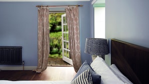 Ein Schlafzimmer mit Bett, Nachttisch, Lampe und Blick auf einen Garten durch ein Fenster mit Vorhängen.