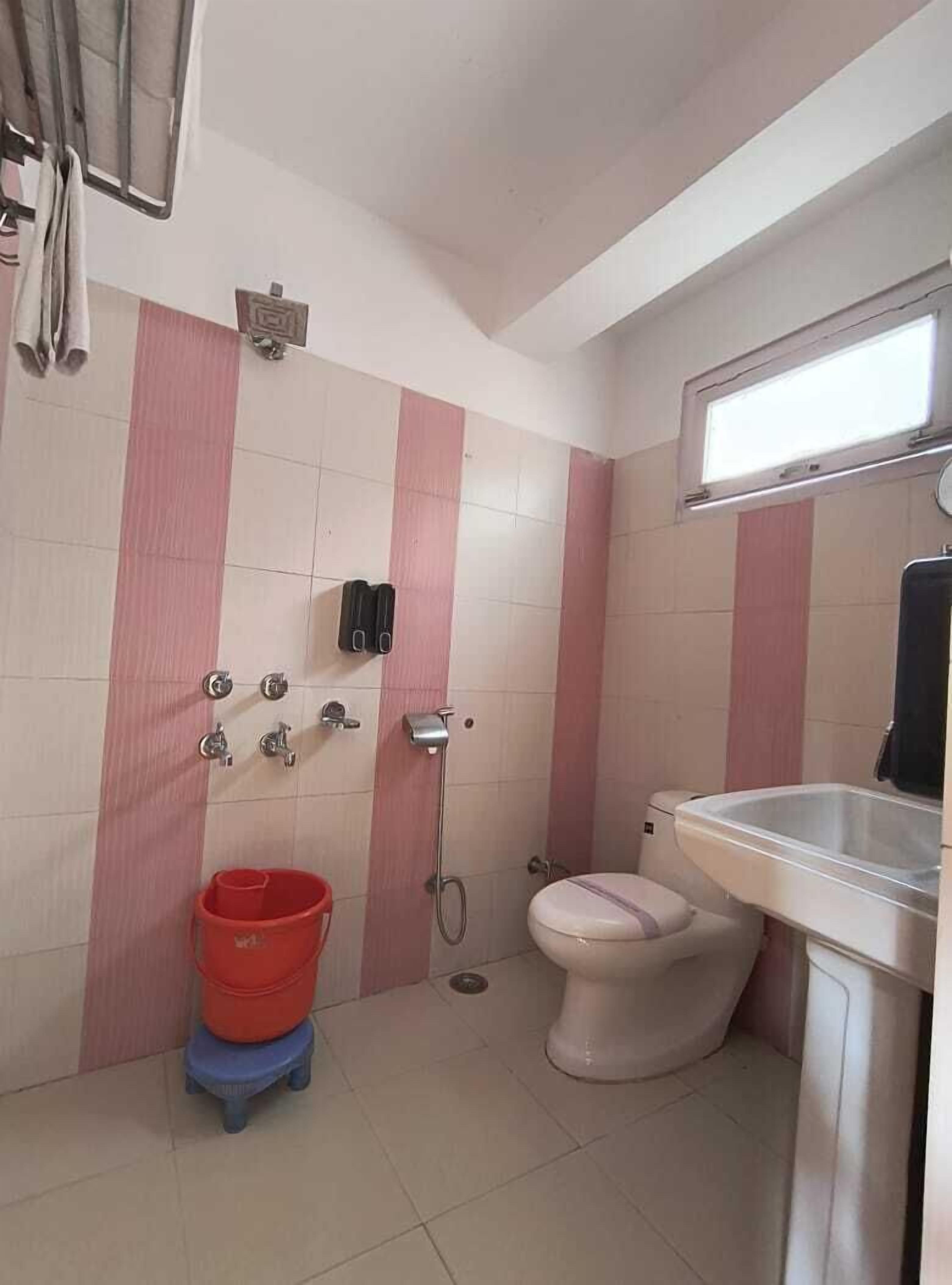 A small bathroom with a toilet, sink, and shower. There is a red bucket and a blue stool.