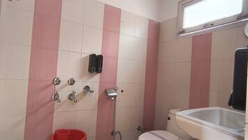 A small bathroom with a toilet, sink, and shower. There is a red bucket and a blue stool.