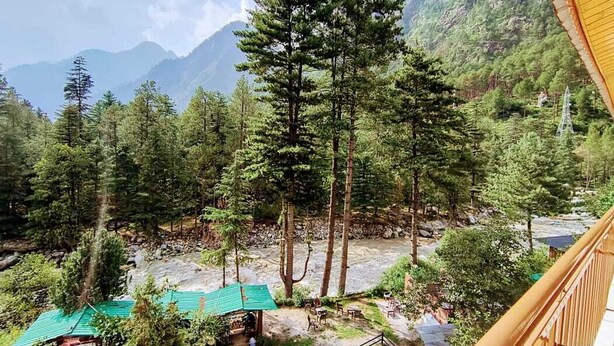 A mountainous landscape with a river, dense forest, and a small outdoor seating area with tables and chairs. - Hotel Purnima River View (Bhuntar)