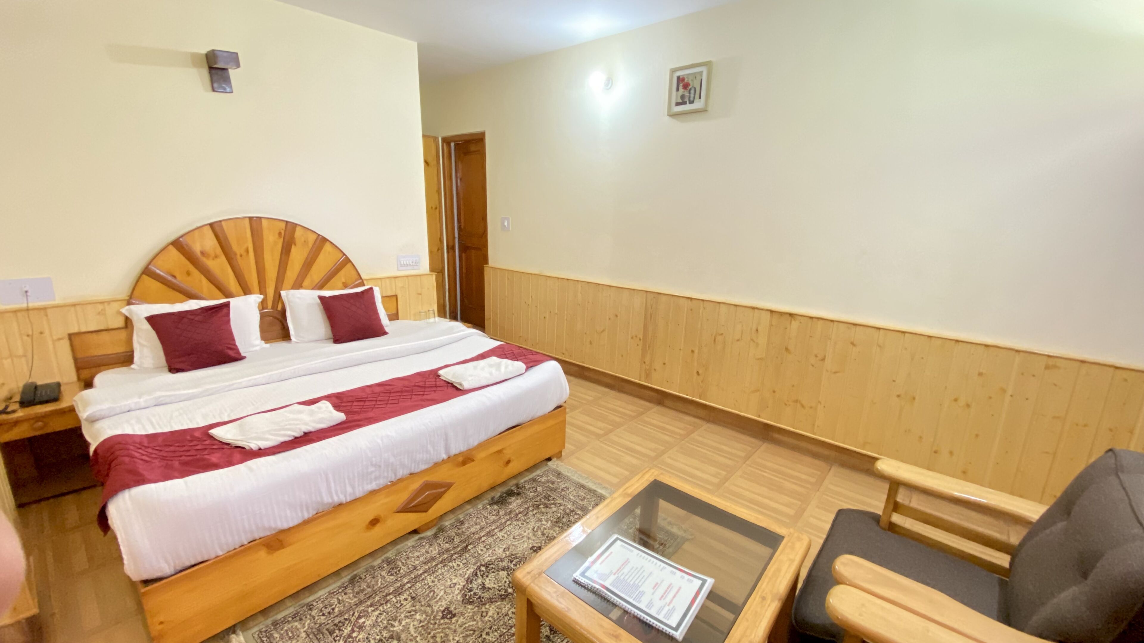 A hotel room with a wooden bed, a small table with a notebook, a chair, and a wooden floor.
