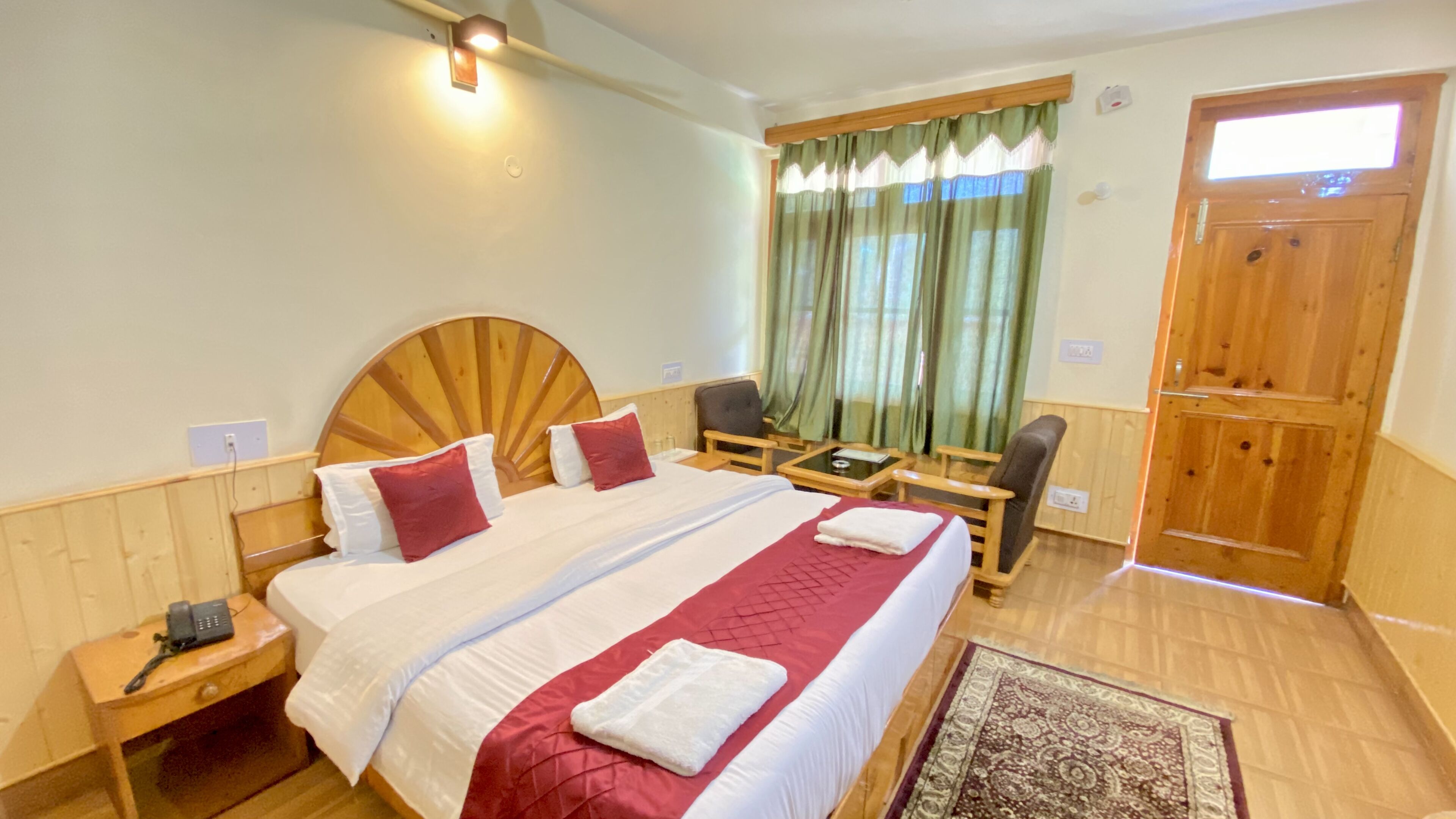 A hotel room with a large bed, two bedside tables, a desk, a chair, a window with curtains, and a wooden door.